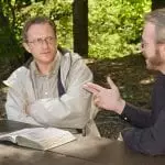 Two men with an open Bible outdoors, one is skeptically listening to the other: